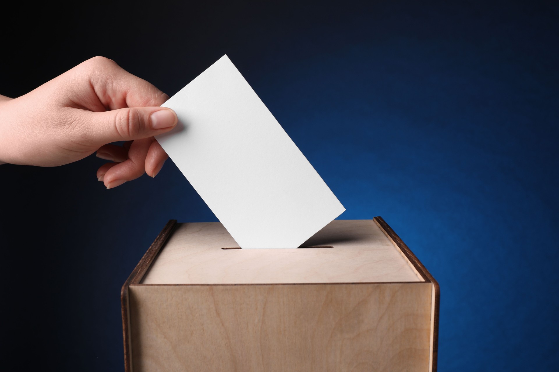 Woman putting her vote into ballot box on dark blue background, closeup