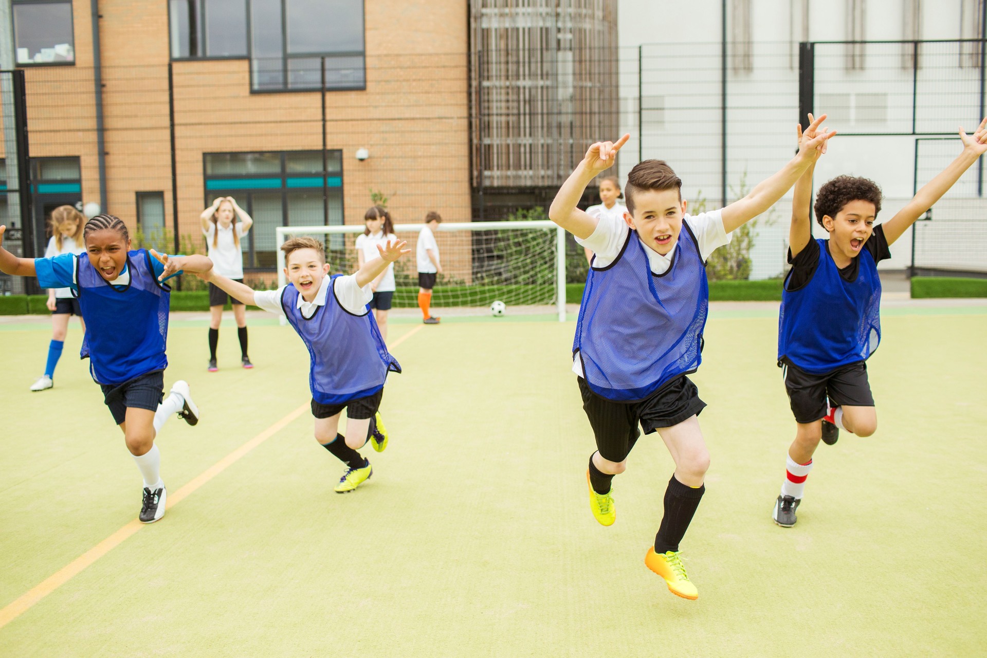 Schoolboys wearing sport uniforms running with arms raised in soccer field in front of school